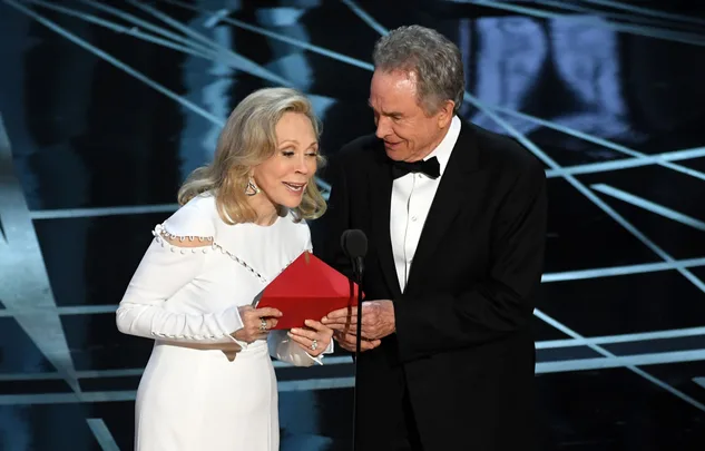 Two people on stage at an awards ceremony, holding a red envelope, smiling and looking at each other.