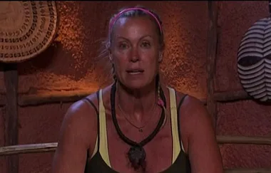 Woman in a tank top and necklace sits on a tribal-themed set with decorative masks on the wall.