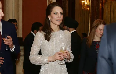 A woman in a sparkling dress holds a glass at a formal event with other attendees in the background.