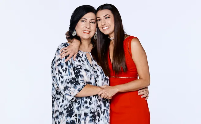 Two women smiling, one in a patterned dress and the other in a red dress, pose against a plain background.