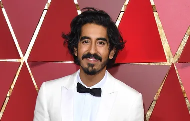 A man in a white tuxedo with a black bow tie stands in front of a red geometric-patterned backdrop, smiling.