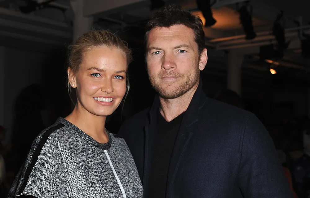 A smiling couple poses indoors; the woman wears a gray top, the man a dark coat.