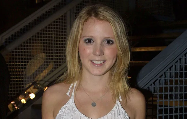 A young woman with long blonde hair smiles at the camera, standing in front of a staircase with metallic railings.
