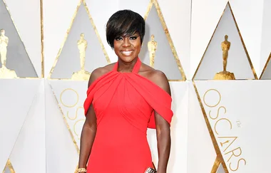 Woman in a red gown poses at the Oscars, with golden statuette decor and the word "Oscars" visible in the background.