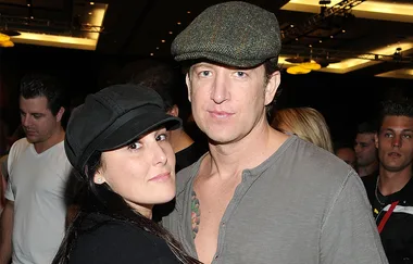 A man and woman wearing caps pose together at an indoor event with a crowd in the background.