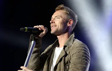 Singer performing on stage, holding a microphone, wearing a brown jacket and white shirt.