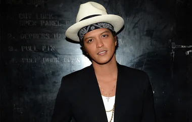 A person wearing a white hat and black blazer stands against a dark textured wall.