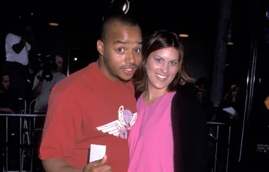 Two people smiling at an event, posing together; the man is wearing a red shirt, the woman a pink top with black cardigan.