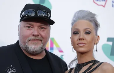 Two individuals posing on the red carpet at an event, with one wearing a cap and the other a dark evening gown.