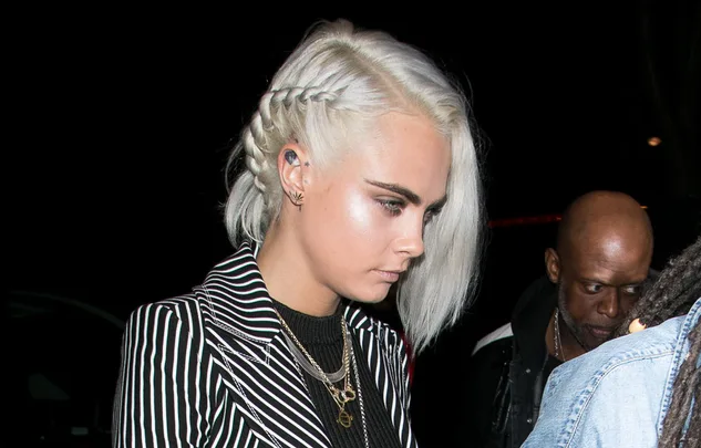 A person with braided silver hair wearing a striped blazer and layered necklaces at a nighttime event.