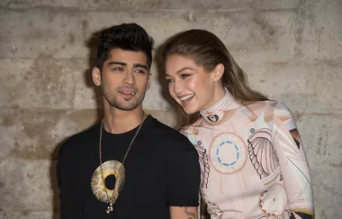 A man in a black shirt stands beside a woman in a patterned outfit, smiling together in front of a stone wall.
