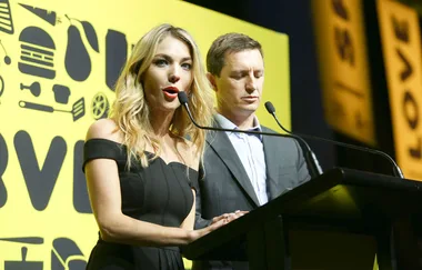 Two people speaking at a podium with a yellow background featuring abstract icons.