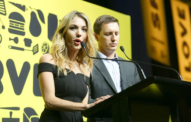 Two people speaking at a podium with a yellow background featuring abstract icons.