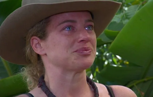 A person wearing a hat, looking emotional with tears, in an outdoor setting with green leaves in the background.
