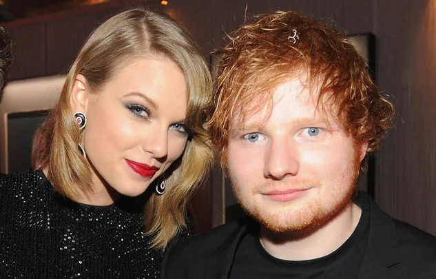 A woman in a black dress and earrings stands smiling next to a man with red hair and a beard, both indoors.