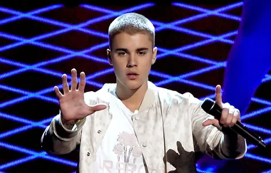 Singer performing on stage while holding a microphone, wearing a beige jacket and white shirt.