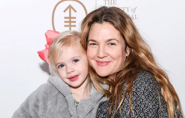 Two smiling people pose together; a child in a gray jacket with a pink bow and an adult with long brown hair.