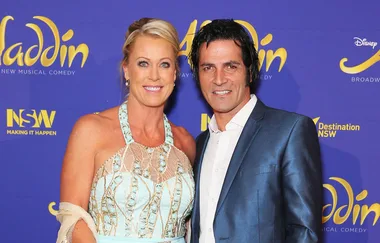 A man and woman posing at the Aladdin musical premiere, smiling in front of a blue and yellow backdrop.
