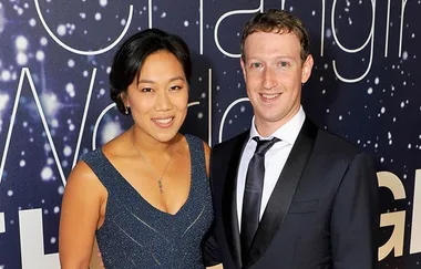 A couple in formal attire smiles on a red carpet with a blue patterned background.