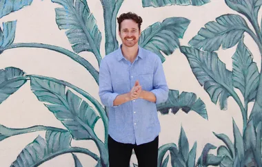 A person smiling, standing in front of a leafy mural, wearing a light blue shirt and dark pants.