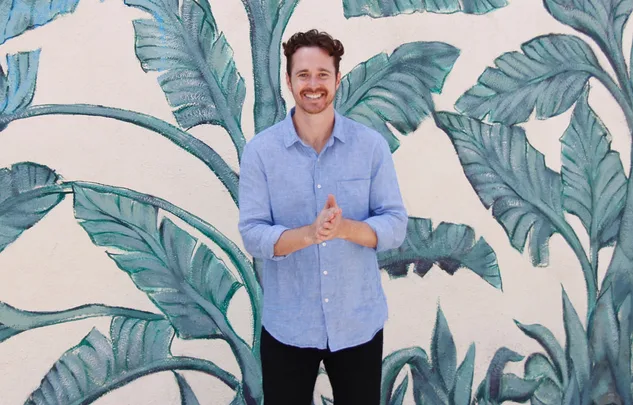 A person smiling, standing in front of a leafy mural, wearing a light blue shirt and dark pants.