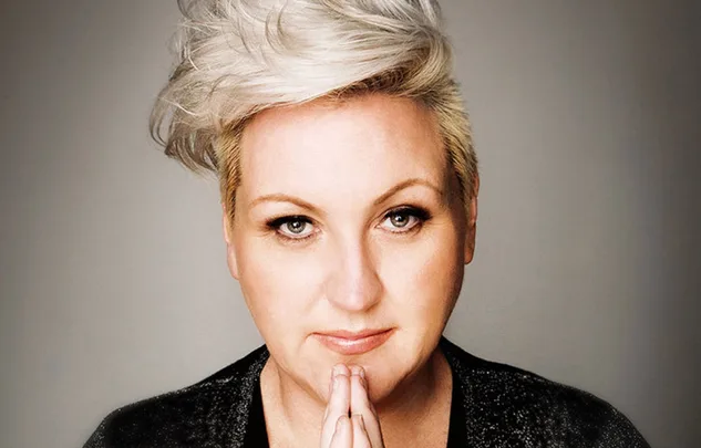 A person with short, stylish platinum blonde hair and a thoughtful expression, hands gently clasped near their chin.