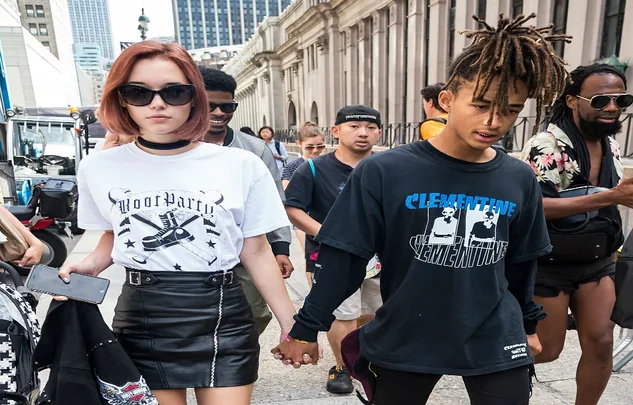 Two individuals walking hand-in-hand on a city street, with surrounding pedestrians and tall buildings.
