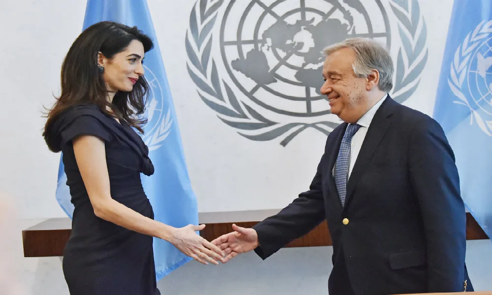Two people shaking hands in front of a United Nations emblem, smiling, with blue UN flags in the background.