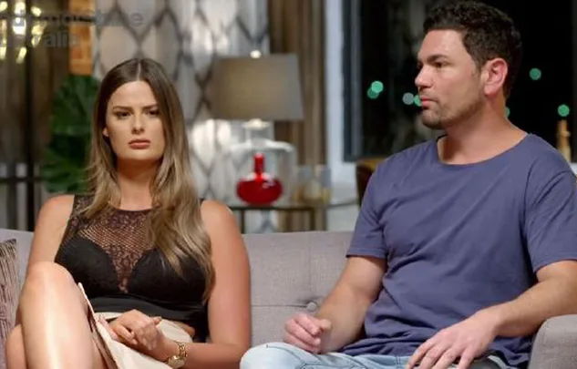Man and woman sitting on a sofa, looking forward in a home setting. Woman wears a black top, man in a blue shirt.