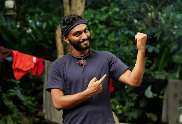 Man in dark shirt and headband playfully flexes arm and points at it in a jungle setting.