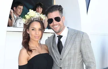 A woman in a black dress with a floral headpiece stands beside a man in a plaid suit and sunglasses, both smiling.
