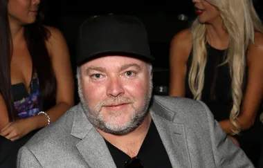 Man in a gray blazer and black cap smiling at an event, with people in the background.