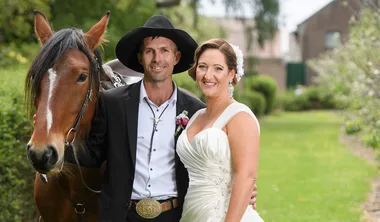 Bride and groom stand smiling with a horse in a garden setting.