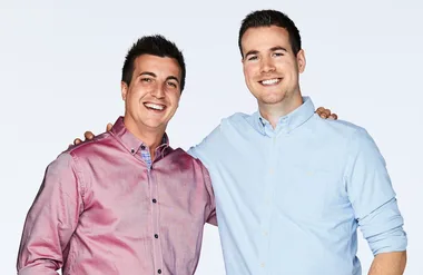 Two men smiling, one in a pink shirt and the other in light blue, with arms around each other against a plain background.