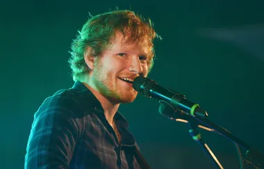 A person with reddish hair singing on stage into a microphone with a green-lit background.