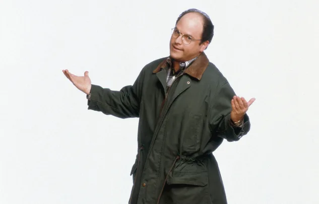 Man in green jacket and glasses, with a puzzled expression and arms slightly raised, against a plain background.