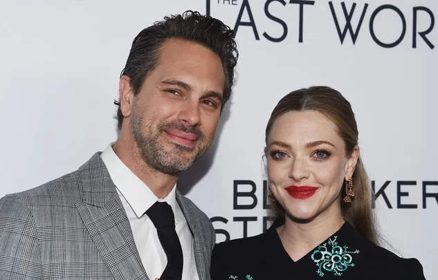Two people smiling at a movie premiere, one in a grey suit and the other in a black dress with embroidery.