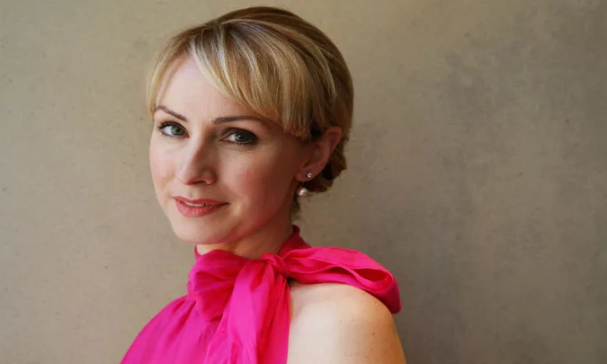A woman with short blonde hair wearing a bright pink dress, looking at the camera, with a neutral background.