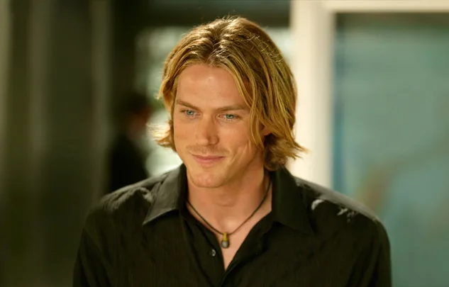Young man with long blonde hair and a necklace, wearing a black shirt, smiling slightly in a bright setting.