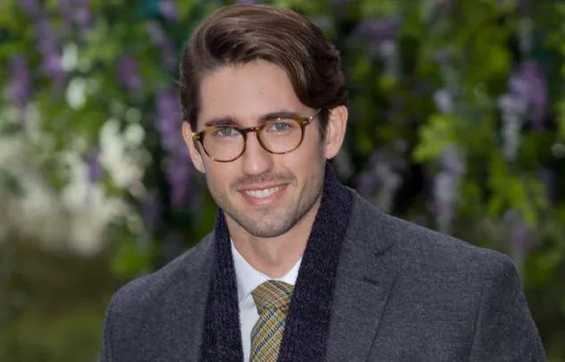Man with glasses smiling, wearing a grey suit and tie, with green foliage in the blurred background.