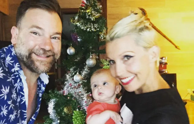 Family posing in front of a decorated Christmas tree, with a baby held by a smiling blonde woman.