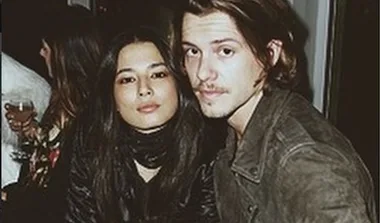 A man and woman sit closely together at a dimly lit gathering, both looking towards the camera.