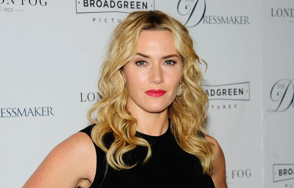 A woman with blonde hair is posing in front of a backdrop with text, wearing a black top and red lipstick.