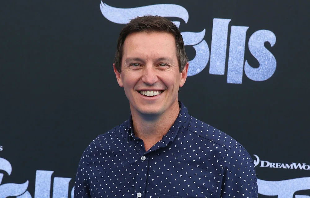 Man smiling at an event, wearing a navy patterned shirt in front of a backdrop with stylized text.