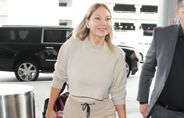 A woman in a beige outfit smiling while walking, with a black SUV in the background.