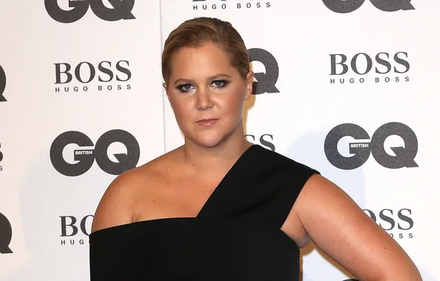 A person posing at the GQ event, wearing a black off-shoulder outfit, standing in front of a branded backdrop.
