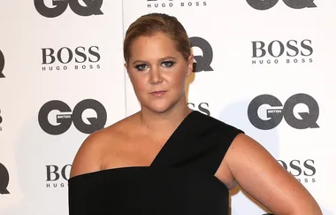 A person posing at the GQ event, wearing a black off-shoulder outfit, standing in front of a branded backdrop.