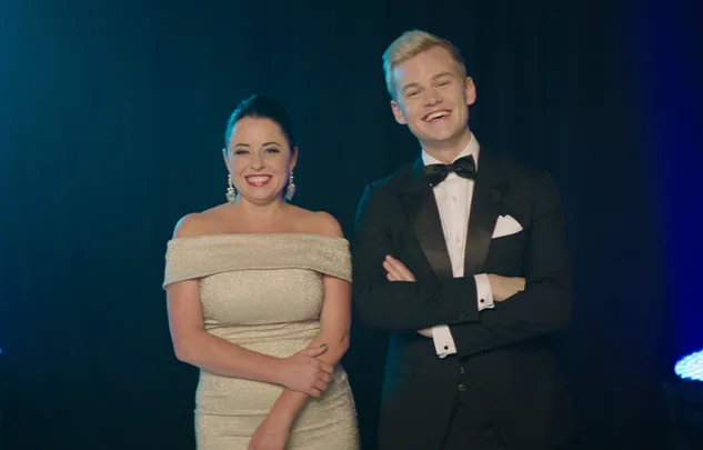 Two people smiling, dressed formally; woman in an off-shoulder gown, man in a tuxedo, standing against a dark background.