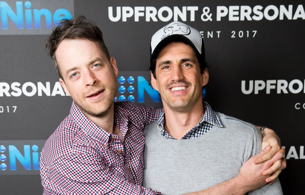 Two men smiling and embracing at a Channel Nine event with the backdrop "UPFRONT & PERSONAL, EVENT 2017."