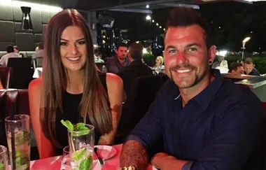 A man and woman smiling at a restaurant table, with drinks and people dining in the background.